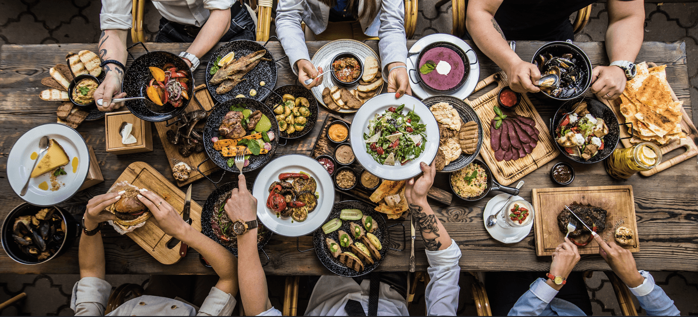 People enjoying a feast on a rustic wooden table with meat, vegetables, soup, and dessert